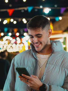 Hombre joven feliz mirando su tel&eacute;fono m&oacute;vil en una calle llena de luces
