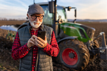 Senior farmer with mobile phone in field with tractor in background