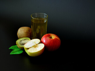 A tall glass of fruit juice on a black background, next to pieces of ripe kiwi and a red apple with leaves.