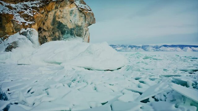 Lake Baikal, flying over broken ice, blue snow hummocks. The camera rises up and reveals a view of the mountain range. A beautiful winter landscape. Baikal, Siberia, Russia. Drone aerial shot 4k
