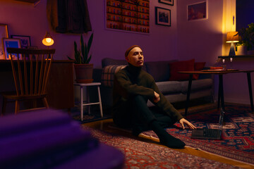A caucasian man wearing a knitted sweater and a beanie sitting on the floor of a living room working on a laptop in a warm light at night.