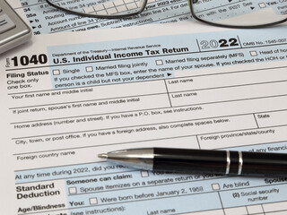 An IRS 1040 tax year 2022 form is shown in 2023, along with an ink pen, calculator, and glasses. The Internal Revenue Service tax filing deadline in 2023 is scheduled for April 18. - obrazy, fototapety, plakaty