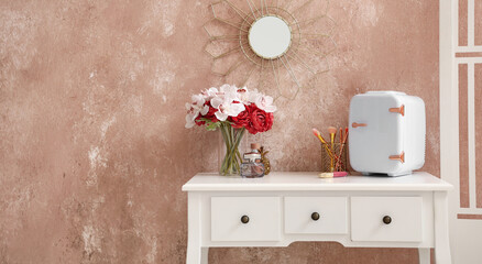 Small cosmetic refrigerator, makeup brushes, perfumes and vase with flowers on table near grunge wall