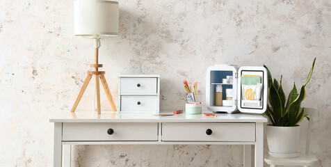 Open cosmetic refrigerator with products and lamp on table near light wall in room