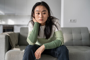 Sad lonely young Asian woman having depression symptoms, sitting on sofa at home looking into distance, upset unhappy female worrying about money or family problems, selective focus. Mental disorders