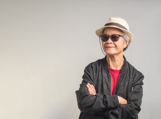 Beautiful elegant elderly woman in a black jacket wearing sunglasses and a hat with a smile while standing with arms crossed on a gray background
