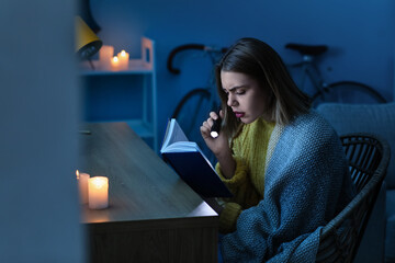 Young woman reading book with flashlight at home during blackout