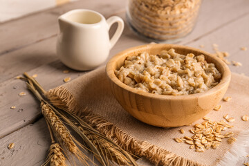Bowl of tasty oatmeal, pitcher with milk, scattered flakes and wheat ears on wooden background