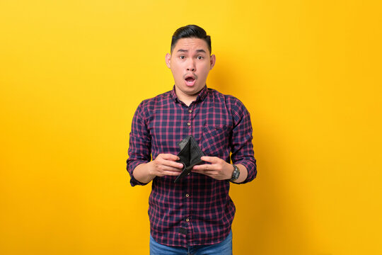 Shocked Young Asian Man Showing Empty Wallet And Looking At Camera Isolated Over Yellow Background. Poverty Concept