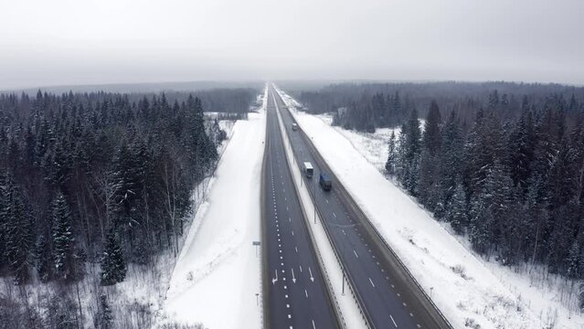 Top view flying a drone back along a road with several trucks passing through a forest covered in snow. The tops of snow-covered trees and the roofs of moving trucks top view