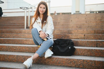 Portrait of a cheerful young girl student with backpack sitting on steps outdoors, reading book