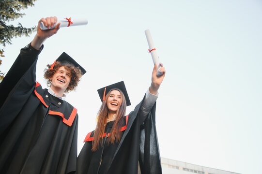 Portrait of happy graduates. Two friends in graduation caps and gowns standing outside university building with other students in background, holding diploma scrolls, and smiling