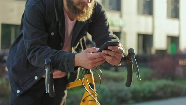 Close up of man with beard standing with yellow bicycle, leaning, holding smartphone. Male wearing casual clothes, texting, scrolling, reading. Concept of urban lifestyle.