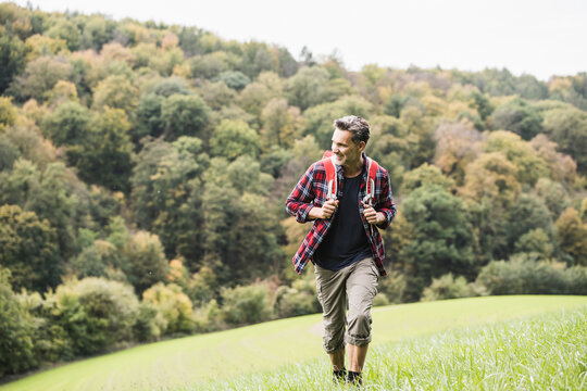 Happy Mature Man With Backpack Walking On Grass By Forest