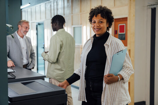 Smiling businesswoman standing by photocopier machine with colleagues standing in background