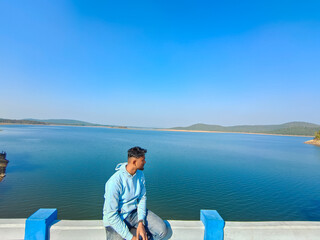 Young man sitting on the bridge and looking at blue river, lake with blue sky scenery. Holiday concept