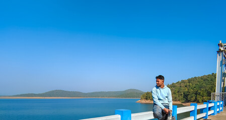 Fototapeta premium Young man sitting on the bridge and looking at blue river landscape view, lake with blue sky scenery. Holiday concept