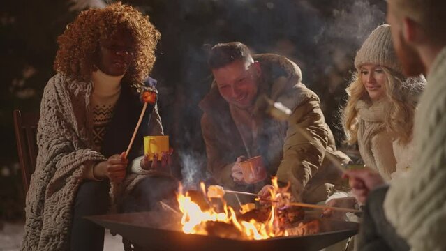 happy male and female friends of different race roasting marshmallows in bonfire in winter night