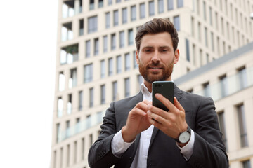 Handsome businessman with smartphone near buildings, space for text