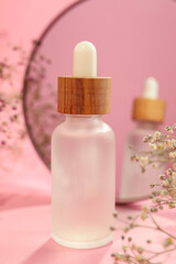 Bottle of face serum and beautiful flowers near mirror on pink background, closeup