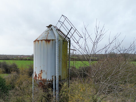 Large, metal grain feed silo showing a  poor state of repair. Surrounding farm buildings are seen belonging to a British dairy farm.