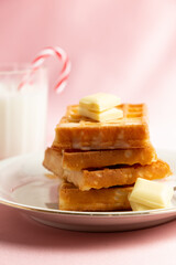 Close up view of a pile of belgian waffles with white chocolate and glass of milk with a candy cane on a pink background