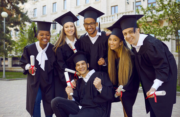 Happy graduates with diplomas posing outside university building. Group portrait of six cheerful joyful mixed race multietnic students and friends in black caps and gowns having fun on graduation day