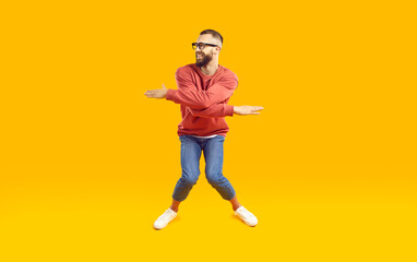 Fototapeta na wymiar Cheerful, funny and energetic man showing humorous joking dance moves on orange background. Full length of young man in stylish casual clothes with funny expression crouching and waving his arms.
