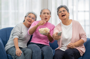 Senior women sitting on couch in living room watching television with excited. Cheerful Asian elderly friends happy excitement watching tv entertainment and eating popcorn. Family relaxation together.