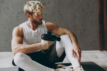Plakat Young serious strong sporty athletic sportsman man wears white tank shirt black shorts warm up percussion massager for body and knee muscles, hand gun, training indoor at gym. Workout sport concept.