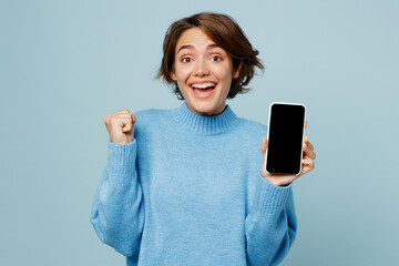 Young smiling happy woman in knitted sweater hold in hand use mobile cell phone with blank screen...