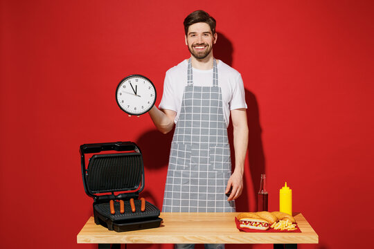 Young housewife housekeeper chef cook baker man in grey apron work at table with grill kitchenware hold in hand clock show time isolated on plain red background studio. Process cooking food concept.