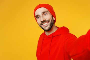 Close up smiling happy fun cheerful satisfied young man in red hoody hat doing selfie shot pov on mobile cell phone isolated on plain yellow color background studio portrait People lifestyle concept