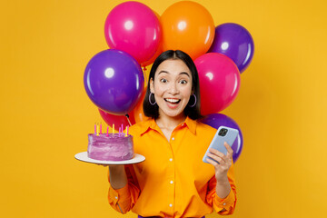 Happy fun surprised young woman wearing casual clothes celebrating near balloons hold cake with candles use mobile cell phone isolated on plain yellow background. Birthday 8 14 holiday party concept. - Powered by Adobe