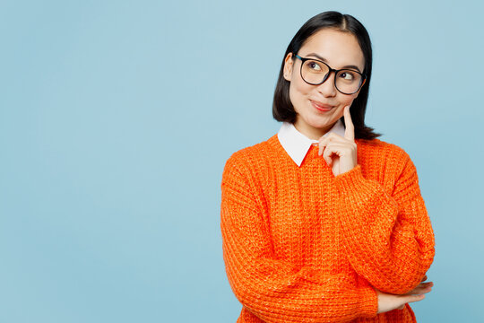 Young minded woman of Asian ethnicity wear orange sweater glasses look aside on workspace area mock up prop up chin isolated on plain pastel light blue cyan background studio People lifestyle concept