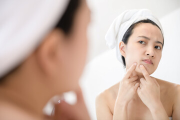 Problem skin. Concerned young asian women popping pimple on cheek while standing near mirror in...