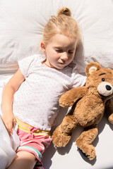 Little girl sleeps hugging a soft toy bear on a white bed. Top view, flat lay