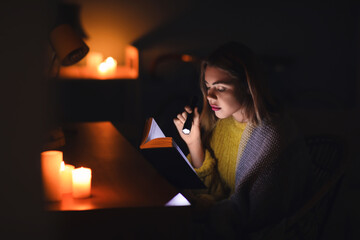 Young woman reading book with flashlight at home during blackout - obrazy, fototapety, plakaty