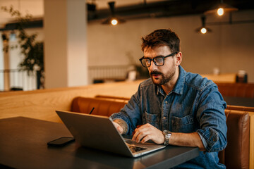 Portrait of a journalist stylish guy writing a story in a workplace in loft styled coworking, casually dressed in a denim shirt, sitting alone.