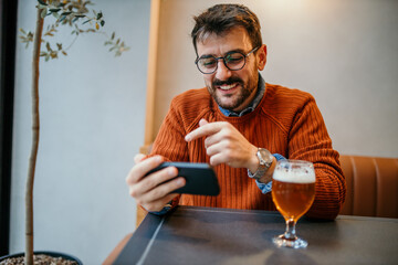 A happy smart casual dressed man sitting in the cafeteria with a beer and smartphone. Businessman watching on smartphone while sitting in a pub restaurant.