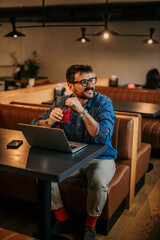 Bearded smiling Caucasian freelancer sitting in a cafe, drinking a fresh smoothie and using a laptop.