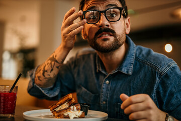 Geek man in a denim shirt sitting in the restaurant and having a sandwich. Portrait of a hungry man with glasses.