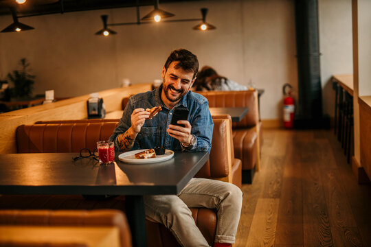 Man in a casual cool outfit sitting in a restaurant, drinking a smoothie, eating a club sandwich, and checking his social media.