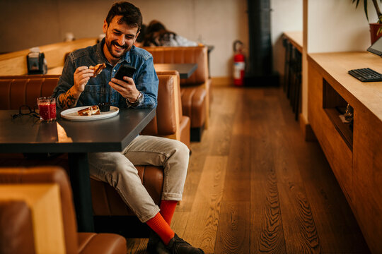 Man in a casual cool outfit sitting in a restaurant, drinking a smoothie, eating a club sandwich, and checking his social media.