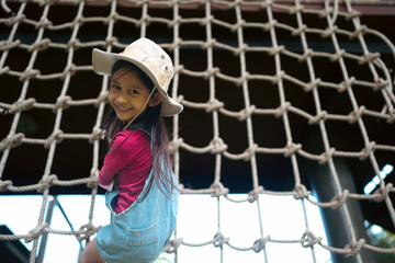 Smiling asian little girl looking to the camera while she is climbing rope net with happy moment. Concept of outdoor activity, play, sport, vacation for kid.