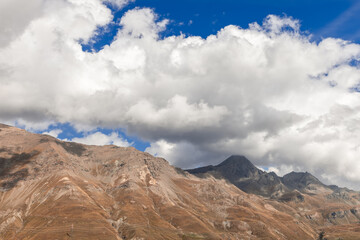 Panorama of autumn yellowed granite slopes of alpine peaks with hiking trails and wires of high-voltage transmission line passing through gorge under heavy white clouds. Aosta Valley, Italy