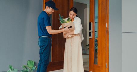 Young Asian delivery man in blue navy uniform knock door home delivery grocery online shopping to...
