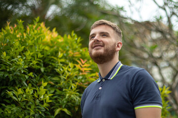 Portrait of a beautiful young bearded man with beard standing on an Ivy wall or in greenery, smiling against from plant, guy relaxing outdoors at sunny summer day. Love nature, eco life