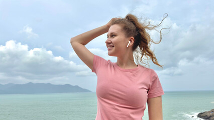 Fototapeta na wymiar Portrait of beautiful girl, young happy woman at sea, ocean background, enjoying summer sunny day, vacation, have fun, listen to music in wireless earphones on smartphone, using phone on mountain