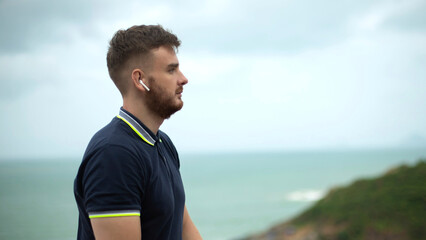 Side portrait of bearded handsome serious pensive thoughtful guy, young man on natural sea, ocean background listening to music, enjoying song in wireless earphones, thinking, dreaming